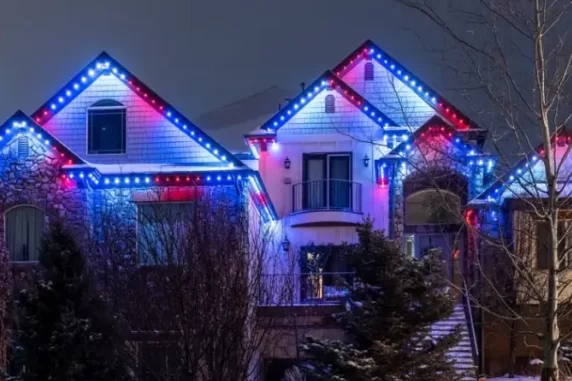 A two-story house is decorated with red, white, and blue lights outlining the roof. Snow covers the yard and trees, and the Trimlight logo appears in the corner of the image.