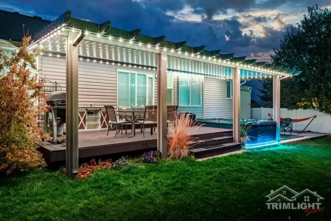 A backyard patio at dusk features a pergola with string lights, outdoor furniture, steps leading down to a grassy lawn, and blue accent lighting along the edge. Trimlight Spirit logo is visible in the corner.
