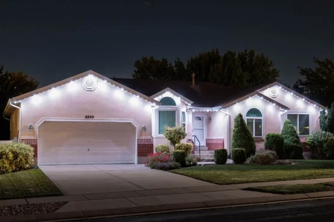 A single-story house at night with white string lights outlining the roof, a two-car garage, neatly trimmed bushes, and a well-kept front yard.