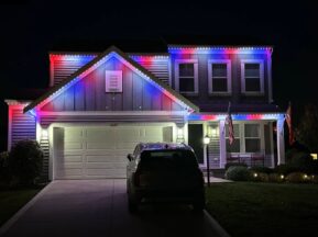 A two-story house at night with red, white, and blue lights outlining the roof. An SUV is parked in the driveway, and two flags are displayed near the porch. The yard is dimly lit.