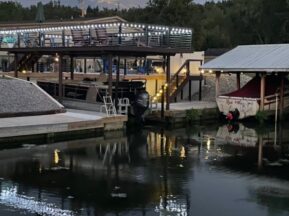 A two-story building with string lights overlooks a calm waterfront at dusk. A covered boat dock with a red and white boat sits to the right, with trees and a full moon visible in the background, reflecting on the water.