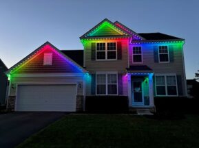 A two-story suburban house at dusk is decorated with colorful LED lights along the roof, glowing in red, yellow, green, and purple, creating a vibrant and festive appearance.