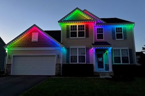 A two-story suburban house at dusk is decorated with colorful LED lights along the roof, glowing in red, yellow, green, and purple, creating a vibrant and festive appearance.