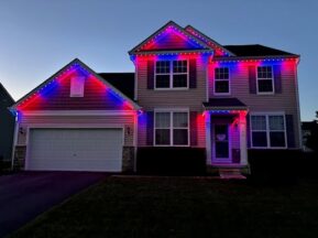 A two-story suburban house is decorated with red, white, and blue lights along the roofline at dusk, giving a festive and patriotic appearance. The house has a double garage and dark shutters.