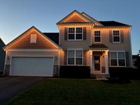 A two-story suburban house with a double garage is shown at dusk. The house is outlined with warm white lights along the roof, illuminating the entrance and creating a cozy, welcoming appearance.