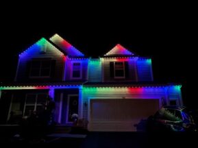 Two-story house at night with white, red, green, and blue lights outlining the roof. A car is parked in the driveway and the garage door is closed. The surroundings are dark.