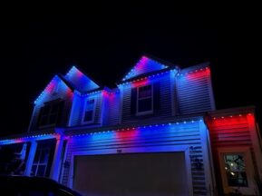 A two-story house at night decorated with red, white, and blue lights along the roofline and garage, creating a patriotic display.