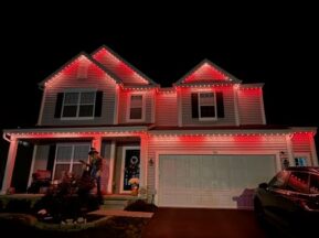 A two-story house at night is decorated with red and white lights along the roofline. The porch is lit, and a Halloween scarecrow stands in the garden near the entrance. A car is parked in the driveway.