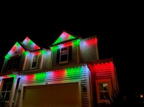 A two-story house decorated with red, green, and white lights along the roofline, glowing brightly against the night sky, creating a festive holiday atmosphere.
