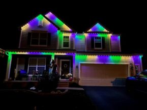 Two-story house at night decorated with bright purple, green, and yellow lights along the roofline. The garage door is closed, and there are Halloween decorations on the front porch.