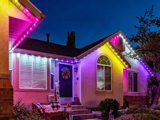 A house at night is decorated with multicolored lights in shades of purple, pink, and white along the roofline. A wreath hangs on the door, and a bench with pillows sits on the front porch. Shrubs and a tree are visible.