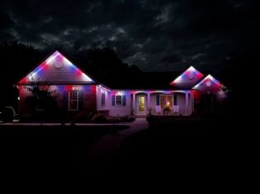 A single-story house at night is decorated with red, white, and blue lights along the roofline, illuminating the front porch and creating a festive atmosphere against a dark, cloudy sky.