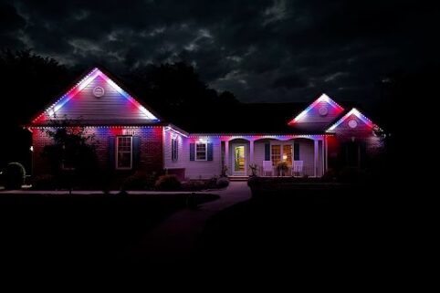 A single-story house at night is decorated with red, white, and blue lights along the roofline, illuminating the front porch and creating a festive atmosphere against a dark, cloudy sky.