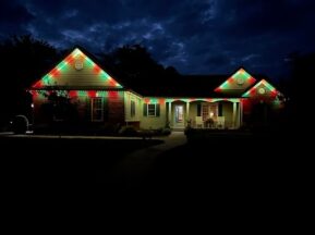 A single-story house at night is decorated with red, green, and white lights along the roofline, creating a festive holiday appearance against a dark, cloudy sky.