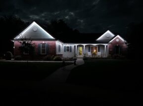 A brick house at night with white lights outlining the roof. The front porch is warmly lit, and two figures, possibly people or animals, stand in the shadowy front yard along a curved walkway.