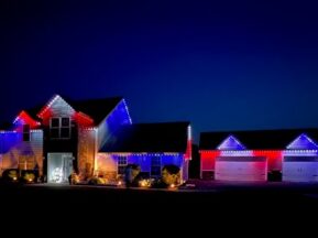A house and three-car garage are decorated with red, white, and blue lights along the rooflines, illuminating the property against a dark night sky. Shrubs and a parked car are visible in the driveway.