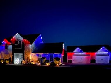 A house and three-car garage are decorated with red, white, and blue lights along the rooflines, illuminating the property against a dark night sky. Shrubs and a parked car are visible in the driveway.