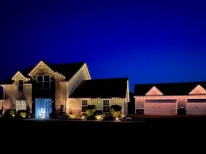 A house and a detached garage are decorated with white string lights along the rooflines, glowing warmly against a deep blue evening sky. Shrubs are lit in the front yard, and a car is parked by the garage.
