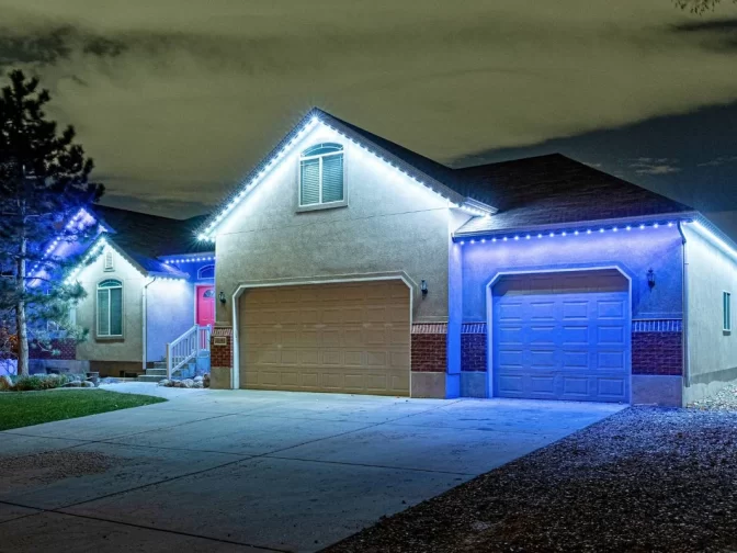 A suburban house at night with a closed two-car garage and single garage, illuminated by bright white and blue string lights along the roof edges, and a red front door visible to the left.