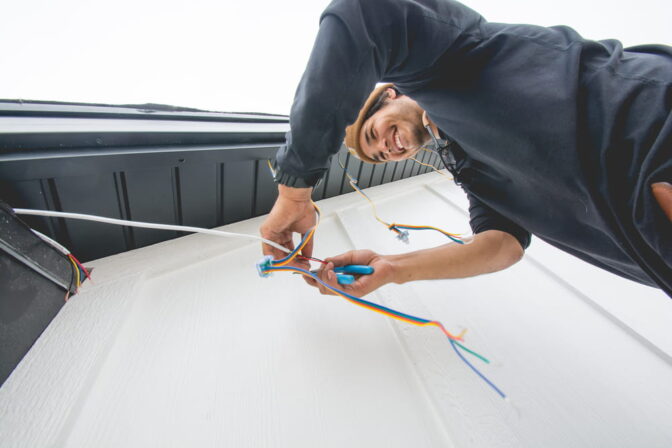 A person smiles while working on electrical wiring outside a building, using tools to connect colorful wires near a white wall and dark roof edge.