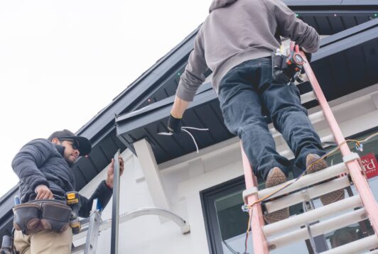 Two workers on ladders install or repair wiring under the roof eaves of a house. One holds tools on his belt, while the other manages wires near the roof edge. Both wear casual work clothes and safety gear.
