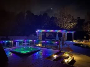 A backyard at night with colorful LED lights outlining a gazebo and deck. There’s a lit hot tub, wooden steps, trees without leaves, and a pool, creating a cozy and vibrant atmosphere under a dark sky.