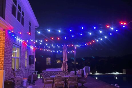 String lights in red, blue, and white hang above an outdoor patio at night, illuminating tables, chairs, a closed umbrella, and a pool next to a house. The scene is festive and calm.