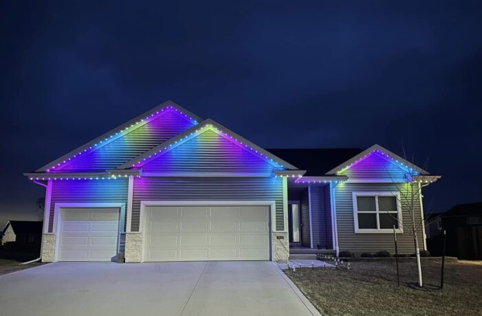 Single-story house at night with colorful LED lights outlining the roof, displaying a gradient of blue, purple, and yellow hues; the house has three garage doors and a large driveway.