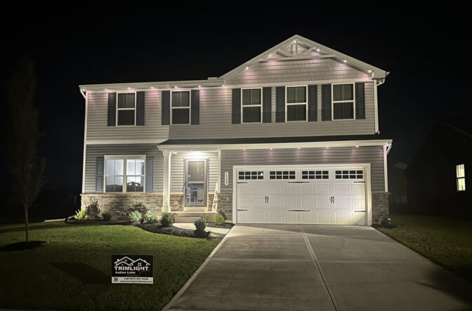 A two-story house at night with lights illuminating the exterior. A Trimlight sign is displayed on the front lawn, and the house has a double garage, neatly trimmed grass, and small shrubs by the entrance.