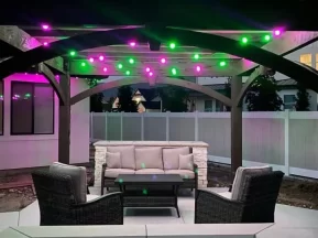 Outdoor patio with wicker chairs, a cushioned sofa, and a table under a wooden pergola. Purple and green string lights are hanging above, with a fire pit in the foreground and a white fence in the background.