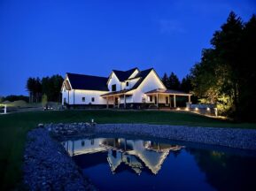 A modern farmhouse with white siding and black roof is lit up at dusk, reflected clearly in a small pond in the foreground, surrounded by grass, trees, and a stone walkway.
