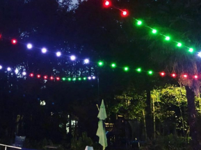 Colorful red, green, and blue string lights are strung over an outdoor pool and chairs at night, surrounded by trees and illuminated by soft lighting. A pool volleyball net is visible in the foreground.
