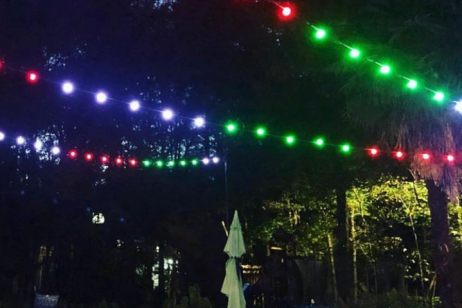 Colorful red, green, and blue string lights are strung over an outdoor pool and chairs at night, surrounded by trees and illuminated by soft lighting. A pool volleyball net is visible in the foreground.