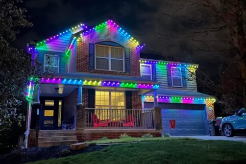 A two-story house decorated with bright, colorful LED lights along the rooflines at night. The lights are in shades of blue, green, pink, and purple, creating a festive look. The yard and driveway are visible in the foreground.