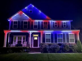 A two-story house at night is decorated with red, white, and blue LED lights outlining the roof and porch, illuminating the front yard and shrubbery.
