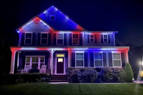 A two-story house at night is decorated with red, white, and blue LED lights outlining the roof and porch, illuminating the front yard and shrubbery.