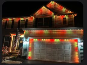 A suburban house is decorated with red, green, and yellow Christmas lights along the roofline and garage. An American flag hangs by the front door, and a small lit tree is in the yard at night.