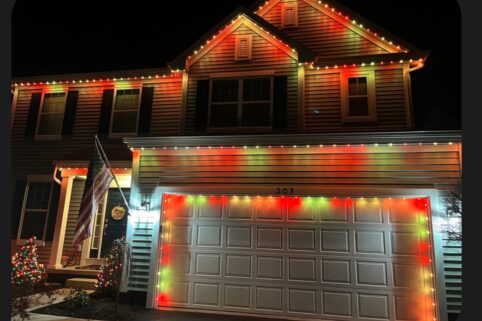 A suburban house is decorated with red, green, and yellow Christmas lights along the roofline and garage. An American flag hangs by the front door, and a small lit tree is in the yard at night.