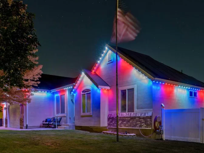 A house at night decorated with red, white, and blue lights along the roofline, with an American flag on a pole in the front yard and a tree and fence visible.