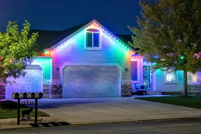 Single-story house at night with colorful LED lights outlining the roof and garage. Trees frame the scene, and four mailboxes are visible at the curb. The driveway and front entrance are well lit.