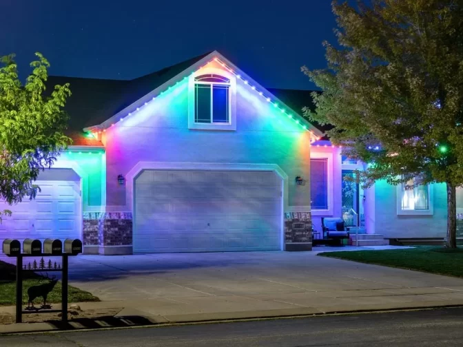 Single-story house at night with colorful LED lights outlining the roof and garage. Trees frame the scene, and four mailboxes are visible at the curb. The driveway and front entrance are well lit.