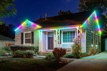 A small house at night with its roofline decorated by colorful, bright LED lights. The yard has bushes and a white bench sits by the front door. The house is well-lit and inviting.