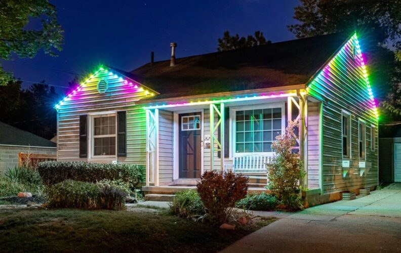 A small house at night with its roofline decorated by colorful, bright LED lights. The yard has bushes and a white bench sits by the front door. The house is well-lit and inviting.