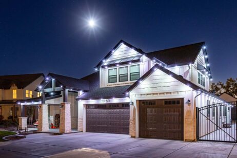 A modern two-story house at night, illuminated by white string lights along the roofline. The moon shines brightly in the clear sky above, casting a glow on the driveway and garage doors.
