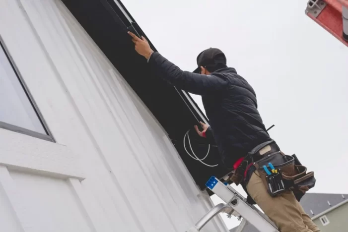 A person standing on a ladder installs or repairs electrical wiring under the eaves of a white house, wearing work gear and a tool belt on their waist.