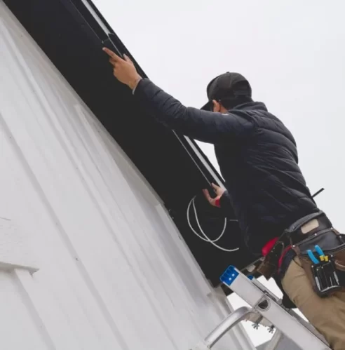 A person standing on a ladder installs or repairs electrical wiring under the eaves of a white house, wearing work gear and a tool belt on their waist.