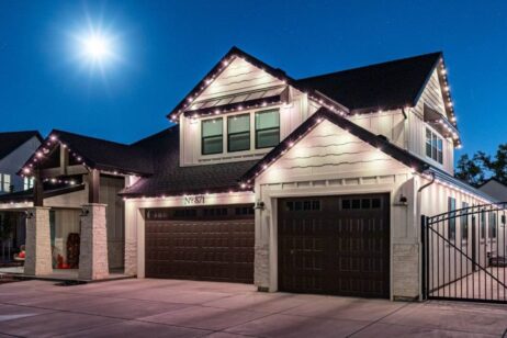 A modern two-story house at night with exterior lights outlining the roof, three dark brown garage doors, and a bright moon shining in the clear sky above.