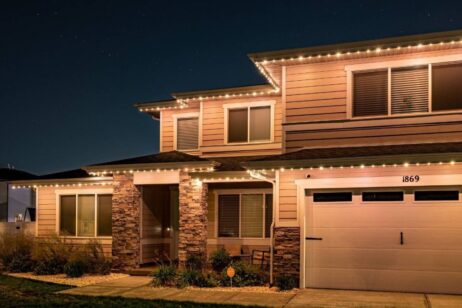 A two-story house with stone accents and beige siding is lit by string lights along the roofline at night. The house number 1869 is visible above the garage.