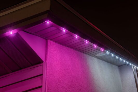 The corner of a house roof at night is lit with purple-pink LED lights along the eaves, casting a colorful glow on the wall, with a few white lights visible farther down the edge.