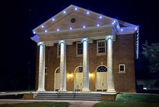 A brick building with four large white columns, labeled Kingsford Office, is illuminated by white lights at night. Three arched double doors are visible, and steps lead up to the entrance.